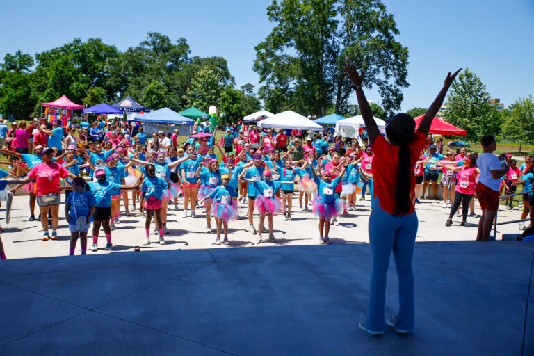 A large group of children and adults participate in a community fitness event at Moncus Park, led by a woman onstage with arms raised, surrounded by colorful outfits and vendor tents.