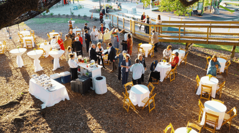Guests enjoy a private outdoor event at Moncus Park with cocktail tables, a bar setup, and shaded seating under oak trees.